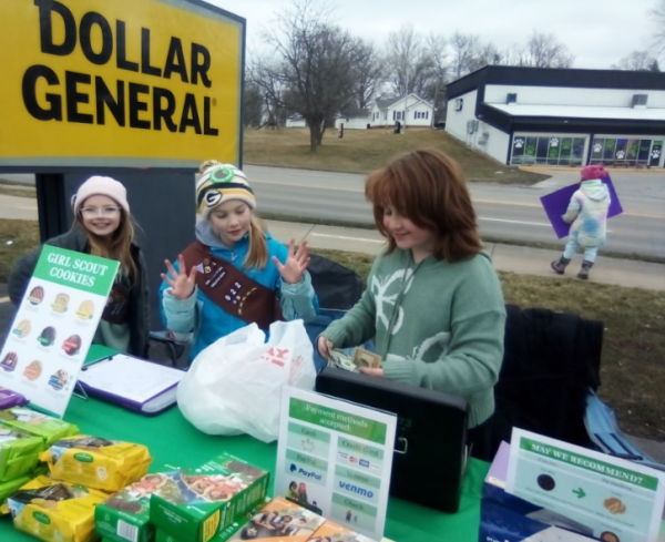 Athena Overturf (left) poses for a photo as Evelyn Hoffman, and Sophie Lowe count money at their cookie booth, while in the background Bree Sammons holds a sign to draw in customers. The cookie booth was on Sunday, Feb. 11, outside of Dollar General. The scouts are trying to earn money for their troop to go camping in the summer. 